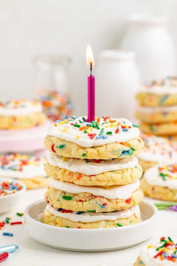 Stack of frosted cake cookies with sprinkles, topped with a lit pink birthday candle on a white plate.