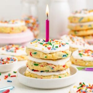 Stack of three frosted birthday cake cookies with sprinkles and a lit purple candle on a plate; cookies in background.