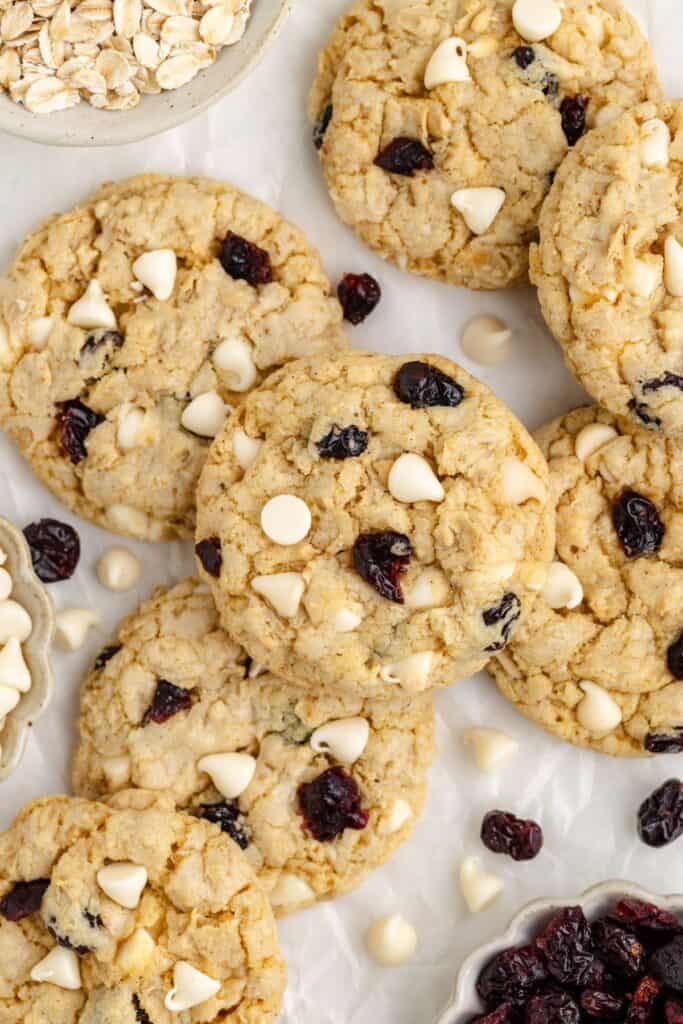 Oatmeal cookies with white chocolate chips and cranberries on a white surface, with small bowls of oats and cranberries.