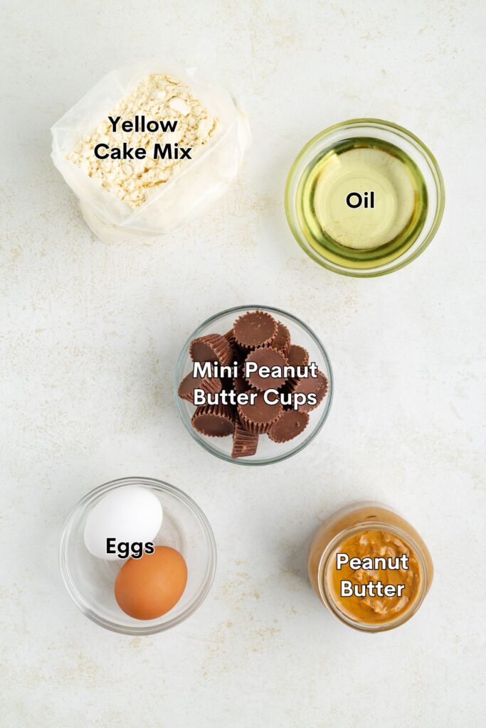 Five baking ingredients—cake mix, oil, mini peanut butter cups, eggs, and peanut butter—on a white surface.