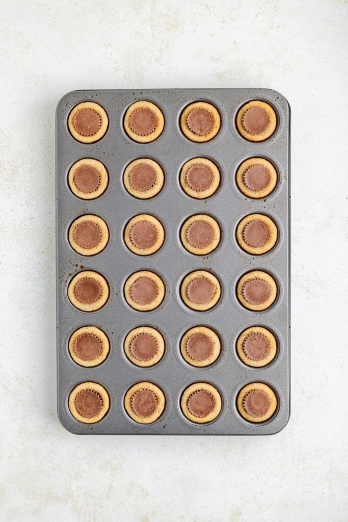 Muffin tin holding 24 peanut butter cup cookies, each with a chocolate center, on a light surface.