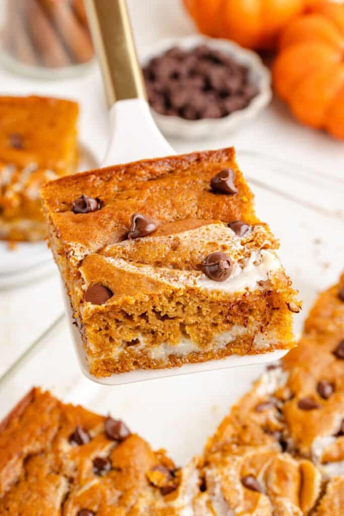 Spatula holding a square of pumpkin earthquake cake with cream swirl over tray; pumpkins in background.