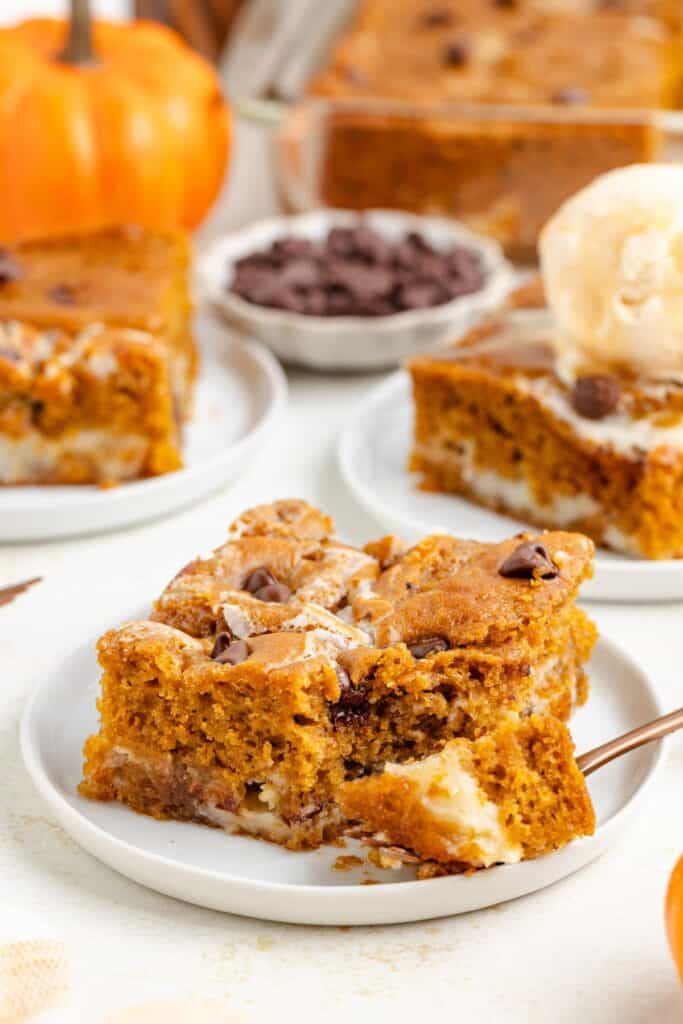 Slice of pumpkin chocolate chip earthquake cake with cream cheese layer on a plate; more cake and chocolate chips behind.