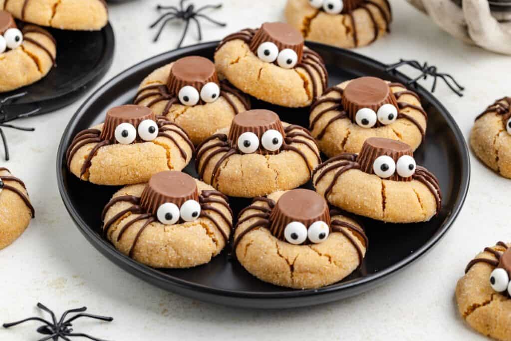 Spider cookies with peanut butter cup bodies and eyes on a black plate, with plastic spider decorations nearby.