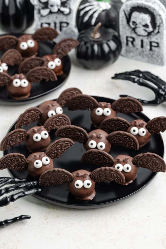 Bat truffles with Oreo wings and candy eyes on black plates, surrounded by Halloween-themed decorations.
