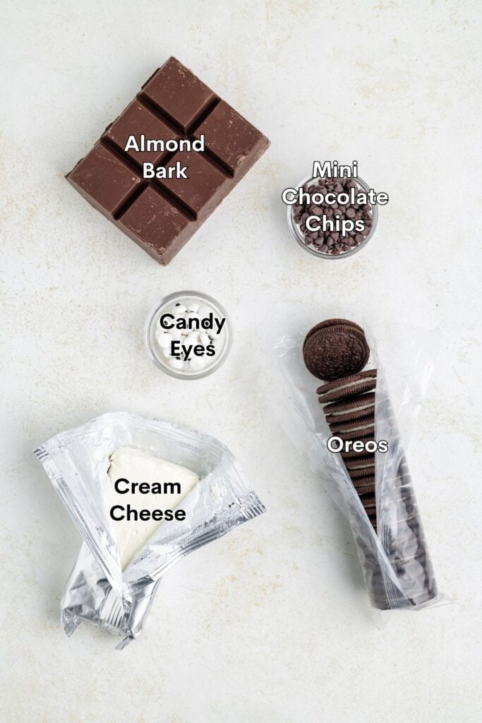 Five labeled ingredients—almond bark, mini chocolate chips, candy eyes, Oreos, and cream cheese—on a white surface.