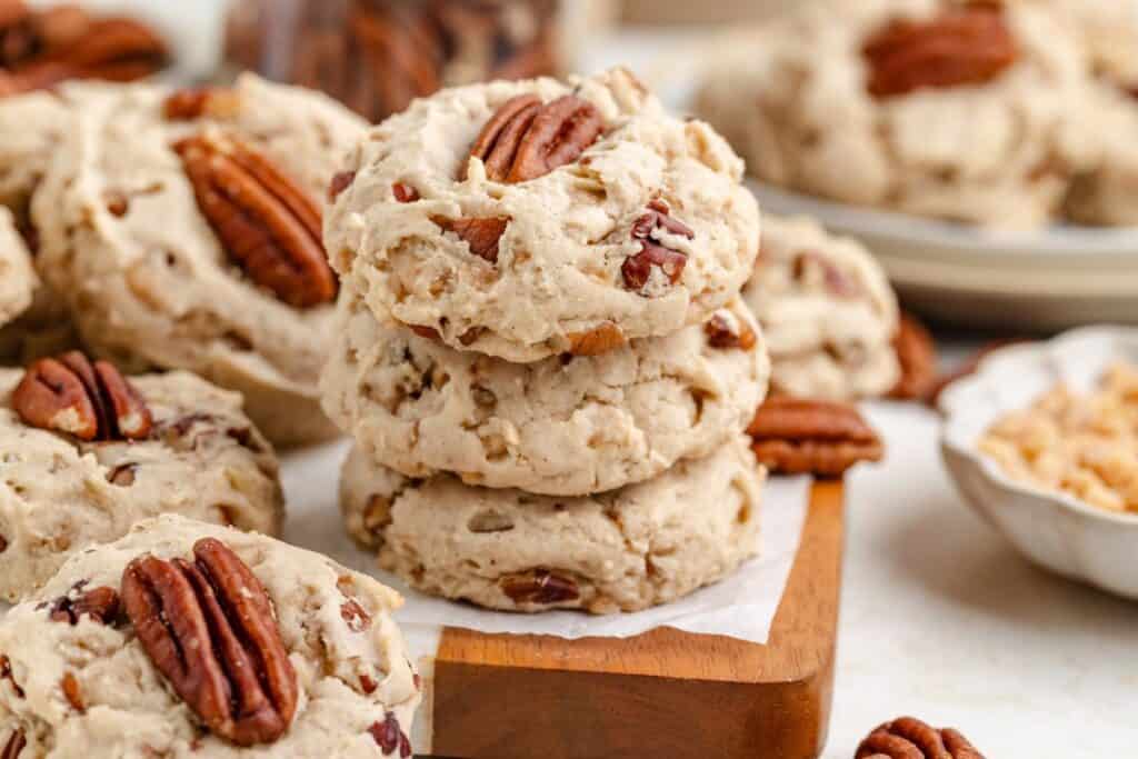Three butter pecan cookies stacked on a wooden board, with scattered cookies and loose pecans around them.