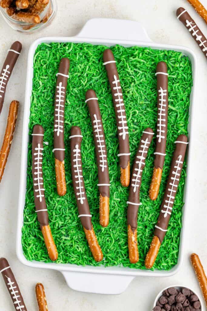Chocolate-dipped pretzel rods decorated like footballs, displayed on green shredded coconut in a white dish.