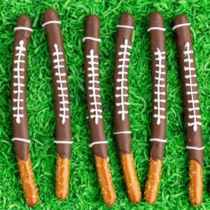 Six chocolate-dipped pretzel sticks decorated as footballs, arranged on a grass-like green surface.