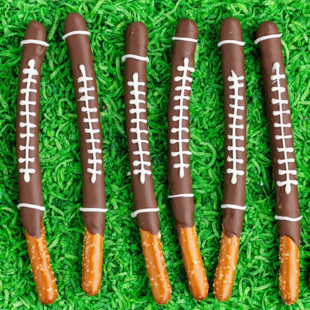Six chocolate-dipped pretzel sticks decorated as footballs, arranged on a grass-like green surface.