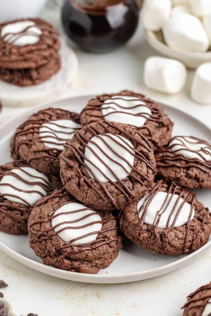 Hot chocolate cookies with marshmallow centers and chocolate drizzle on a plate, with extra cookies in the background.