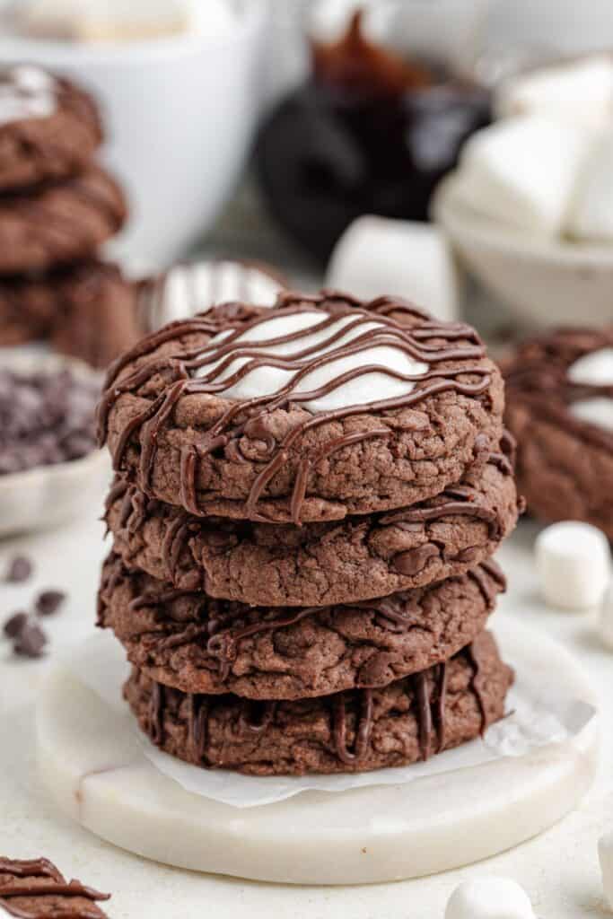 Three chocolate cookies stacked with marshmallow and chocolate drizzle, more cookies and marshmallows behind them.