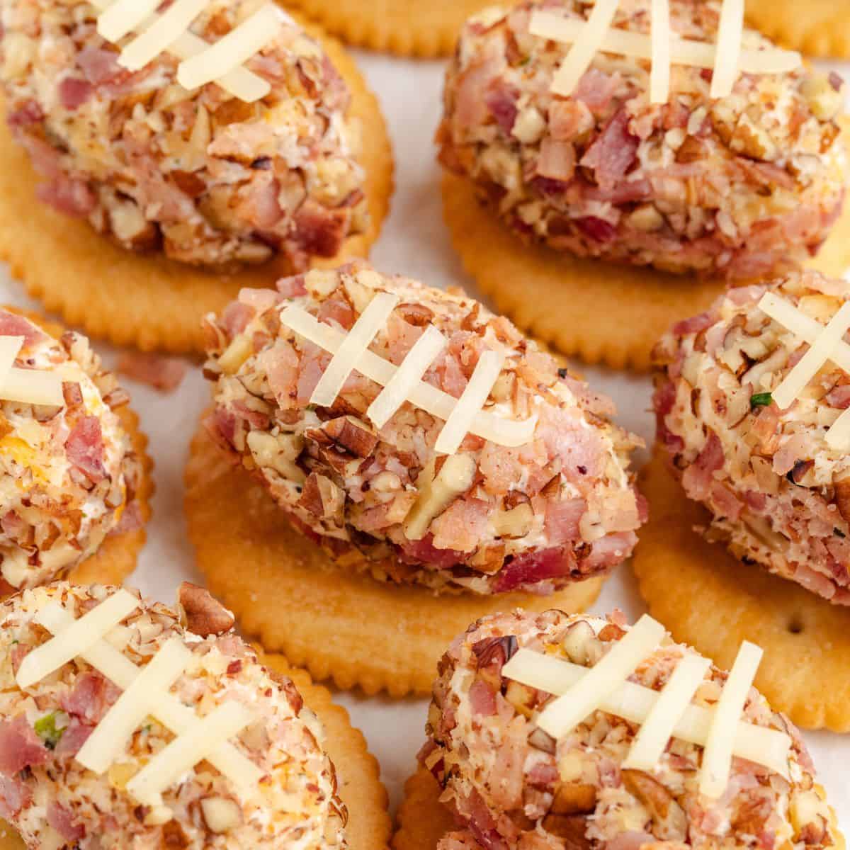 Mini football cheese balls coated in nuts and bacon, topped with cheese strip laces, served individually on round crackers.