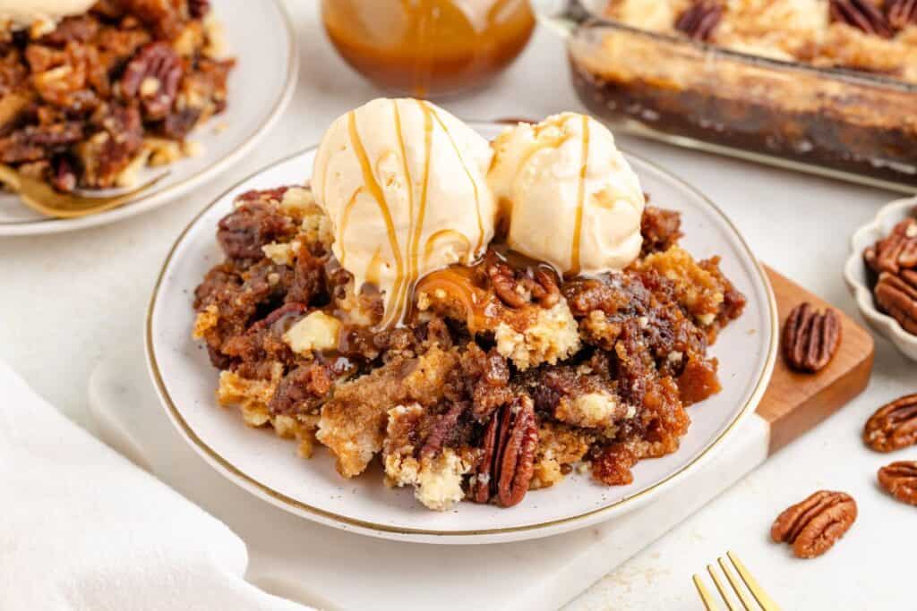 Pecan pie cobbler on a plate, topped with vanilla ice cream, caramel drizzle, and scattered pecans.