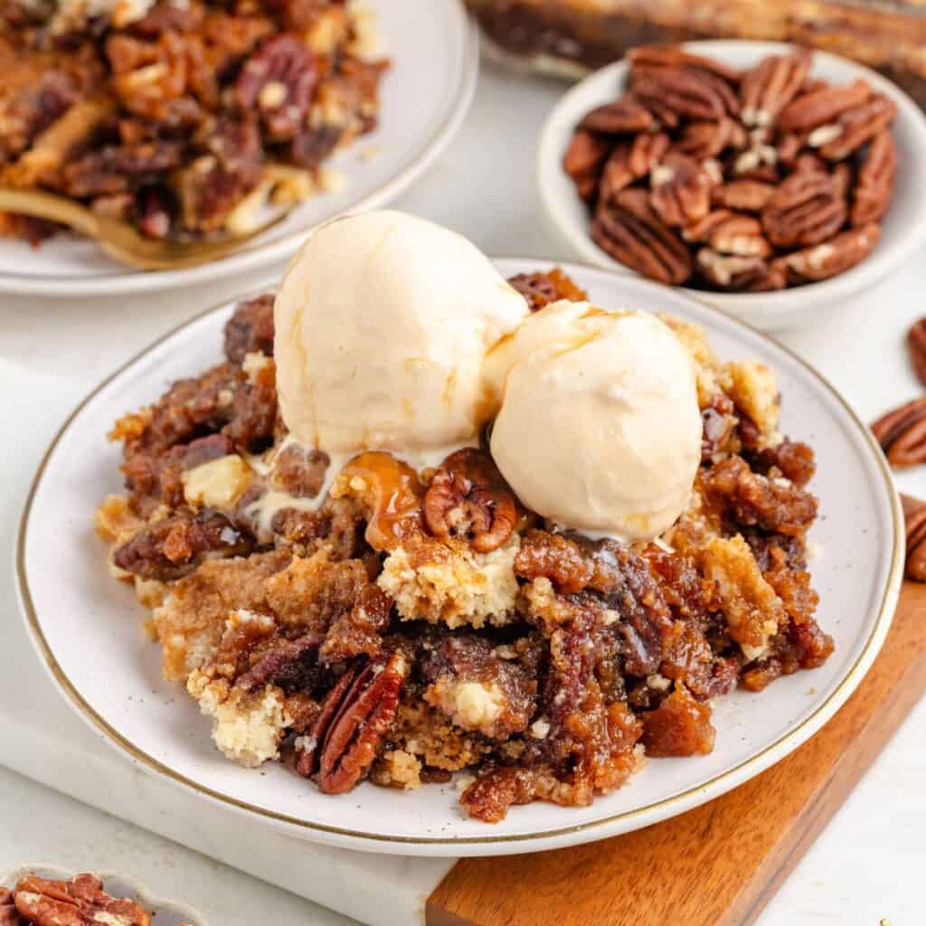 Pecan pie dump cake with two scoops of vanilla ice cream on a plate; whole pecans in the background.