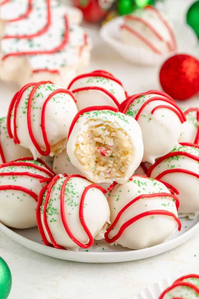 White coated Christmas tree cake truffles with red icing and green sprinkles on a plate; one has a bite revealing the inside.