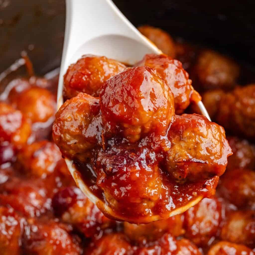A white spoon holds glazed meatballs in thick reddish-brown sauce, with more meatballs behind.
