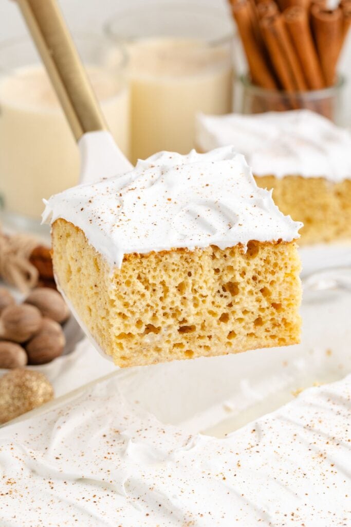 A square eggnog tres leches cake with whipped topping is lifted from a pan; creamy drinks and cinnamon sticks are behind it.
