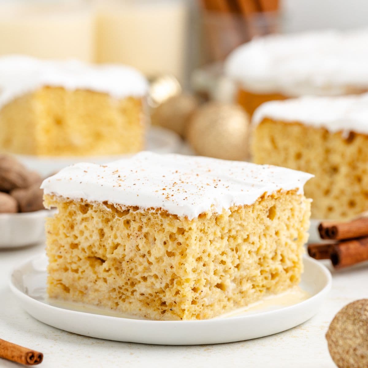 A slice of eggnog tres leches cake on a plate, cinnamon sticks and nuts in the background.