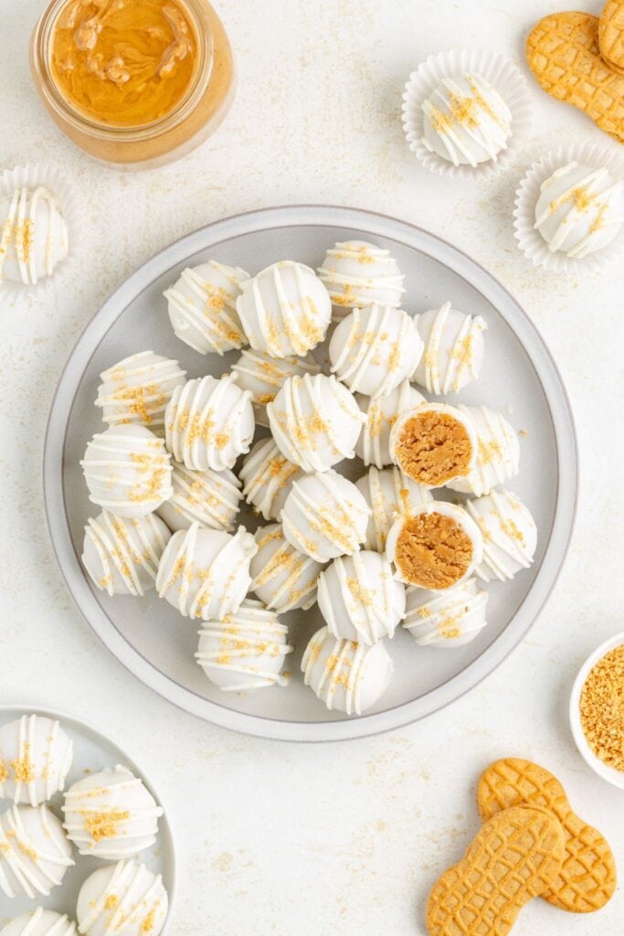 Plate of white chocolate peanut butter cookie truffles, some with crumbs, peanut butter and whole cookies nearby, one truffle cut open.