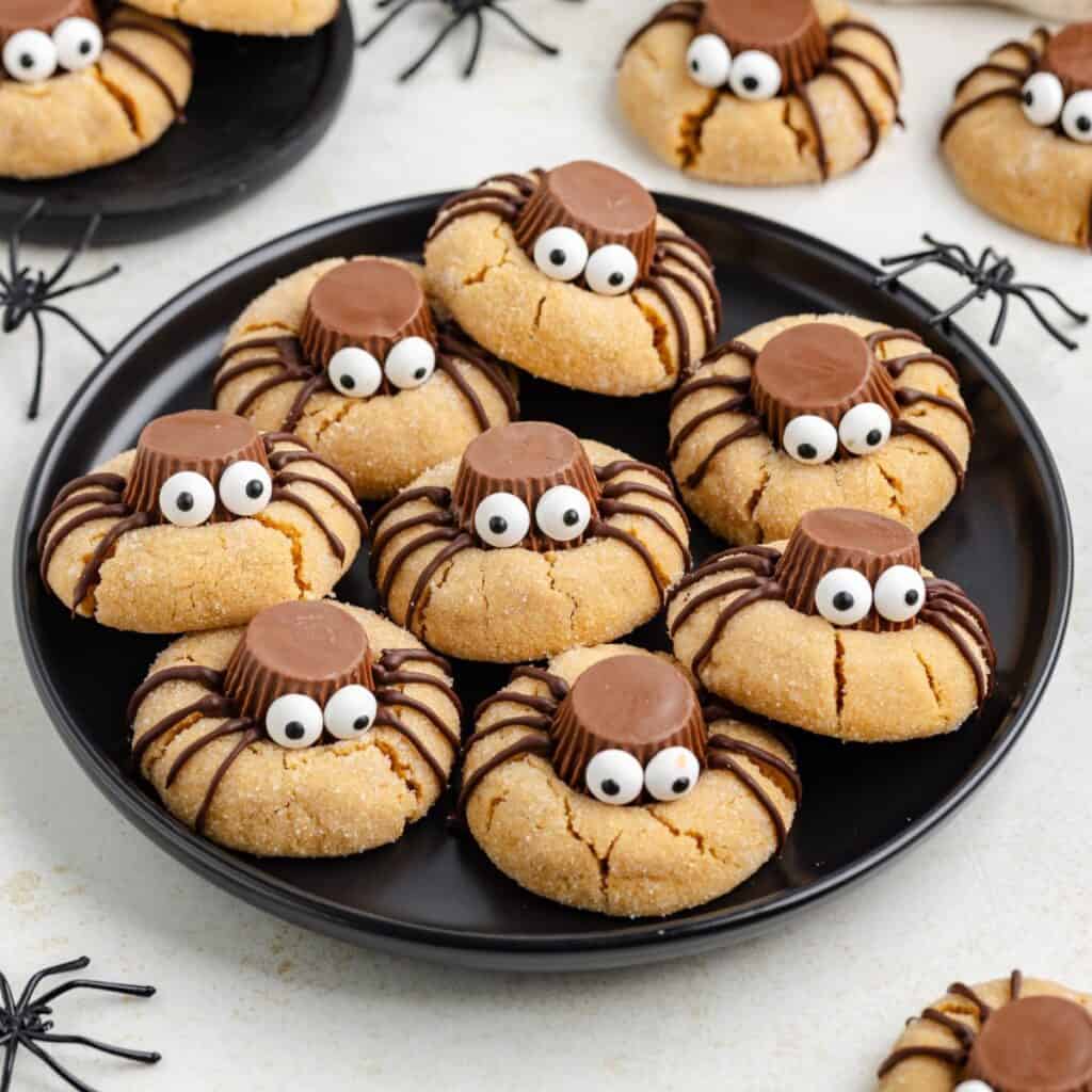 Cookies decorated as spiders on a black plate, with candy eyes and toy spiders around the plate.
