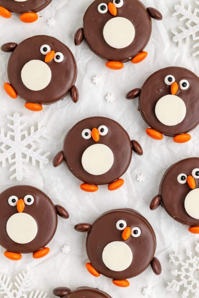 Penguin chocolate cookies with white bellies, candy eyes, orange beaks and feet on parchment with snowflakes.
