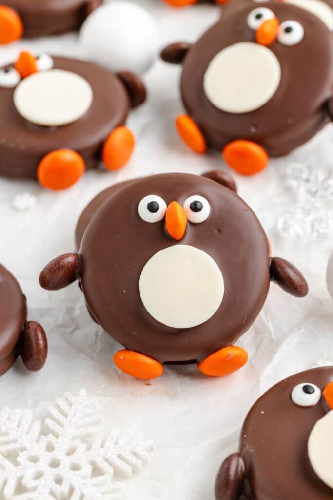 Penguin Oreos with candy eyes, orange beaks and feet, on a white surface with snowflake decor.