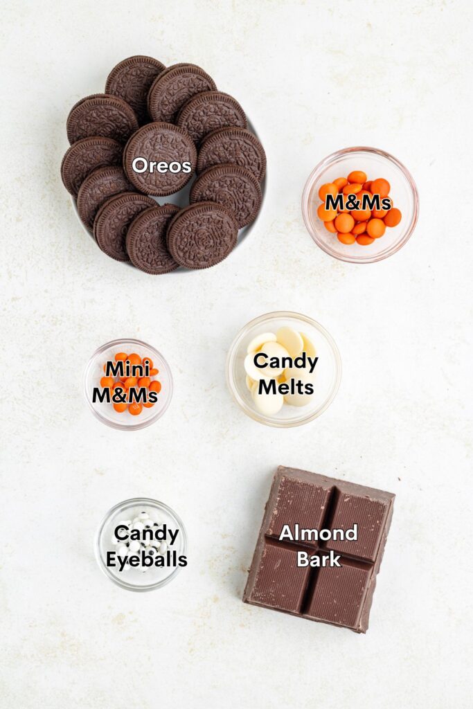 Flat lay of Oreo cookies, orange and mini M&Ms, white candy melts, candy eyeballs, and almond bark on white surface.
