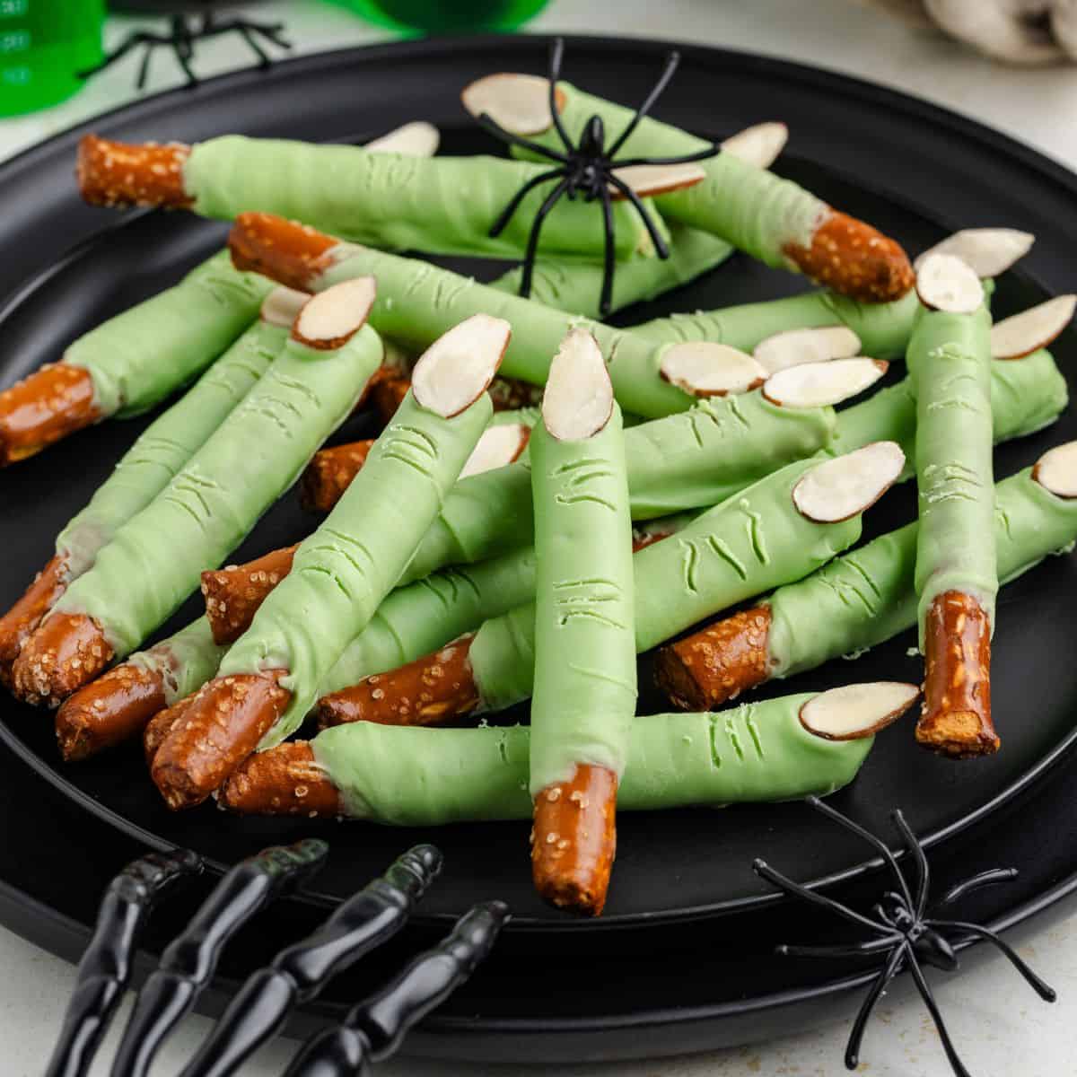 Witch fingers pretzel rods with almond nails on a black plate, plus plastic spiders and skeleton hand.