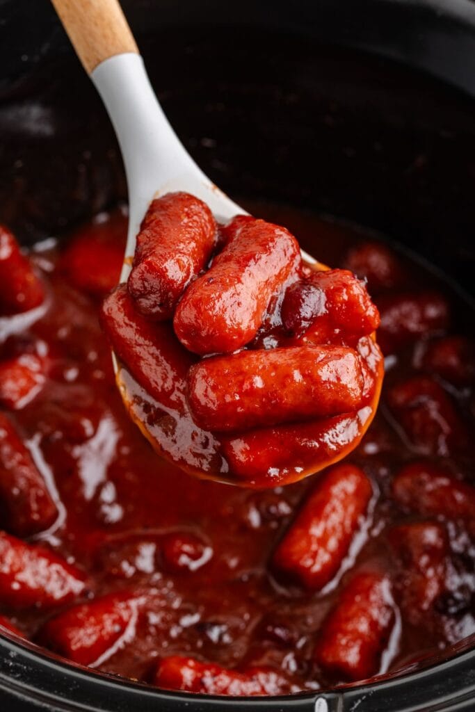 A ladle lifts cranberry cocktail sausages in red sauce above a slow cooker filled with more sausages and sauce.