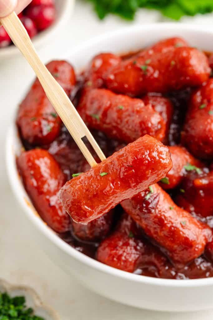 Crockpot Cocktail sausages in red sauce in a bowl, one being lifted by wooden tongs.
