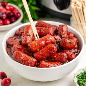 White bowl of slow cooker cranberry little smokies with a toothpick; cranberries and herbs in the background.