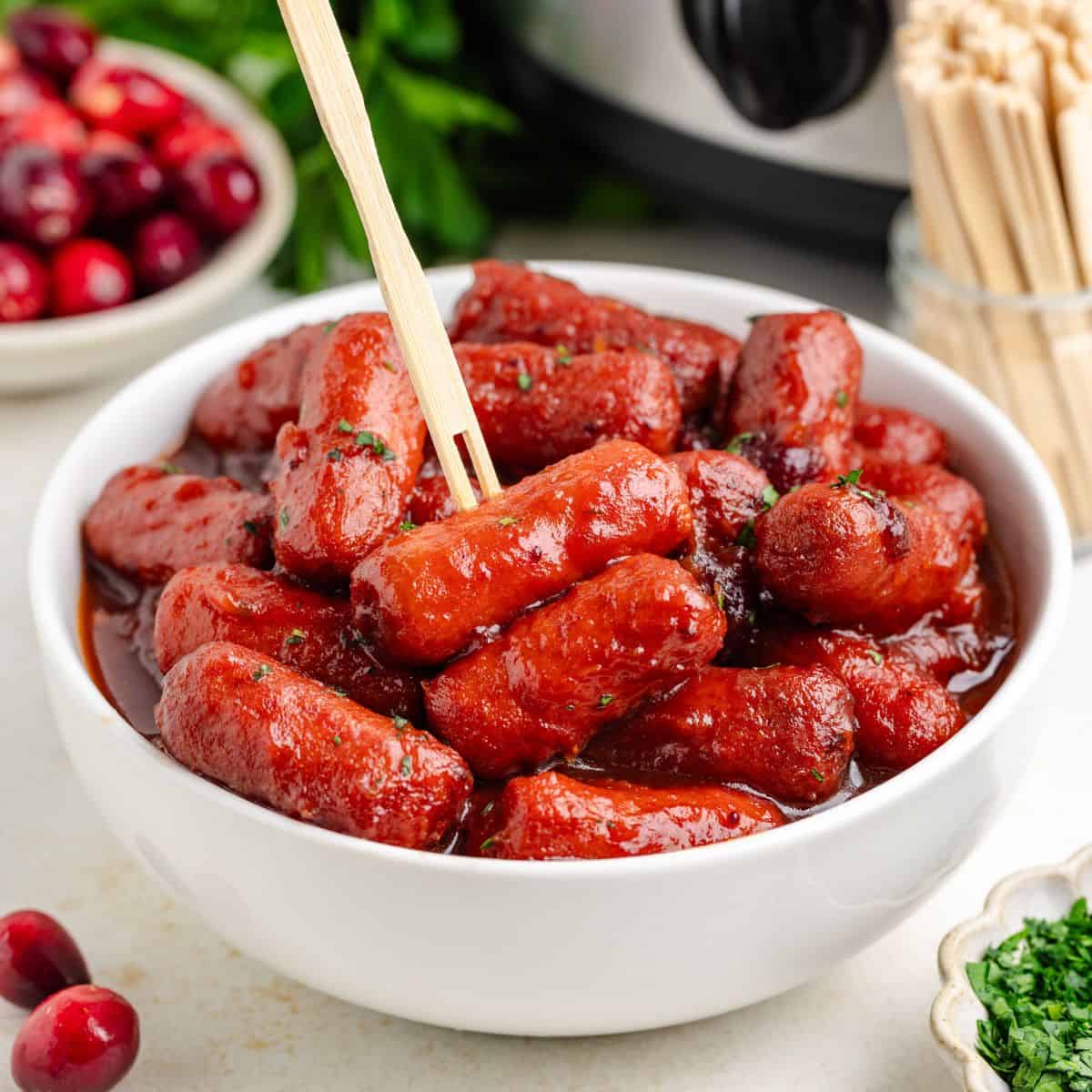 White bowl of slow cooker cranberry little smokies with a toothpick; cranberries and herbs in the background.