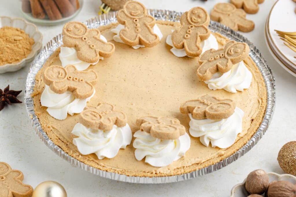 Gingerbread cream pie with, whipped cream dollops, and gingerbread cookies on a white surface.