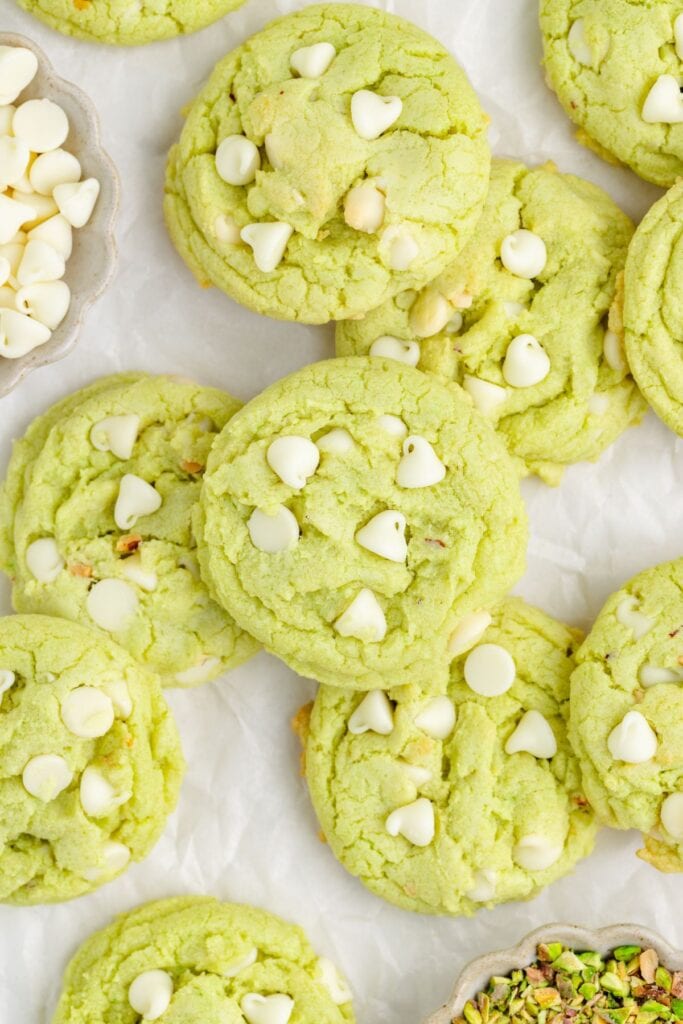 Pistachio Pudding Cookies with white chocolate chips on a white surface, with bowls of extra chips and pistachios nearby.