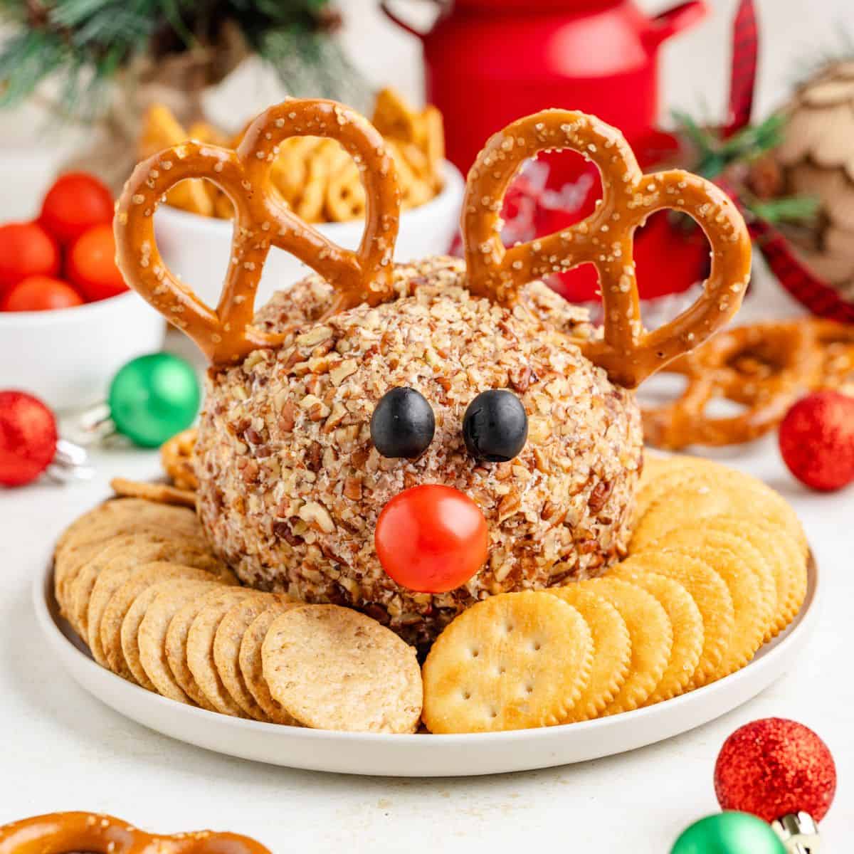 Reindeer cheese ball with pretzel antlers, olive eyes, cherry tomato nose, and crackers on a white plate.