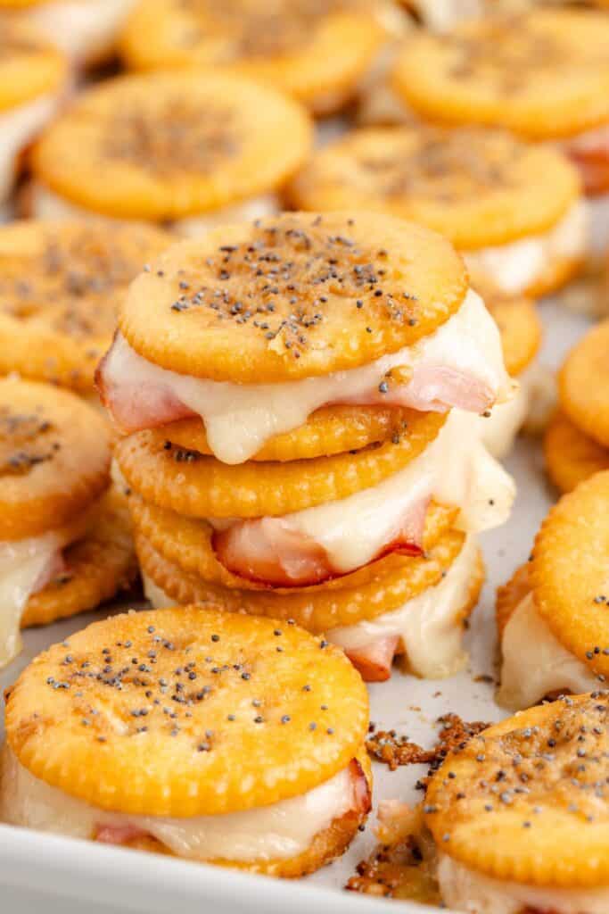 Ritz cracker party sandwiches with melted cheese and ham, topped with poppy seeds, on a baking tray.