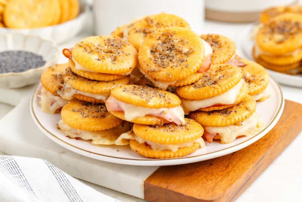 Baked Ritz cracker sandwiches with ham and melted cheese, sprinkled with poppy seeds, served on a wooden board.