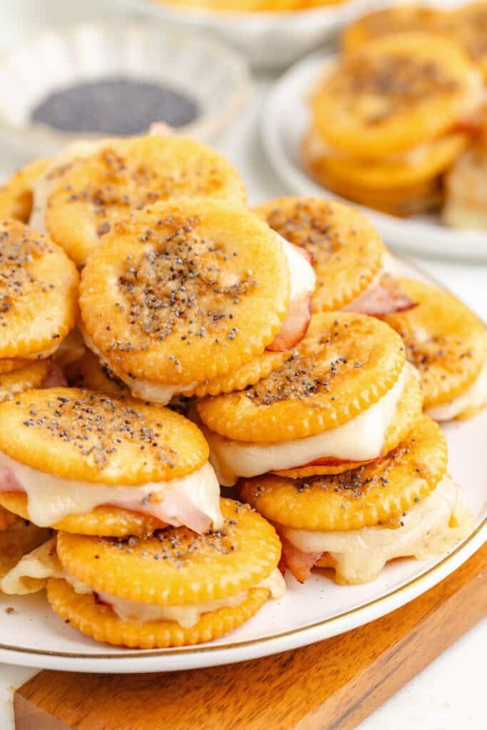 Ritz cheese cracker sandwiches with ham on a plate, next to a dish of black poppy seeds.