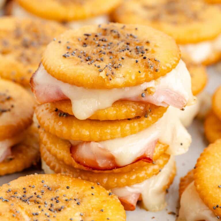 Stack of Ritz cracker party sandwiches with melted cheese, ham, and poppy seeds. More crackers blurred in the background.