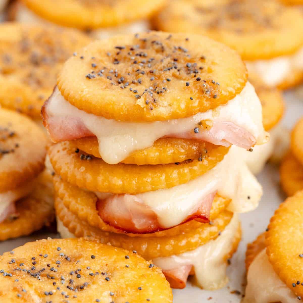 Stack of Ritz cracker party sandwiches with melted cheese, ham, and poppy seeds. More crackers blurred in the background.