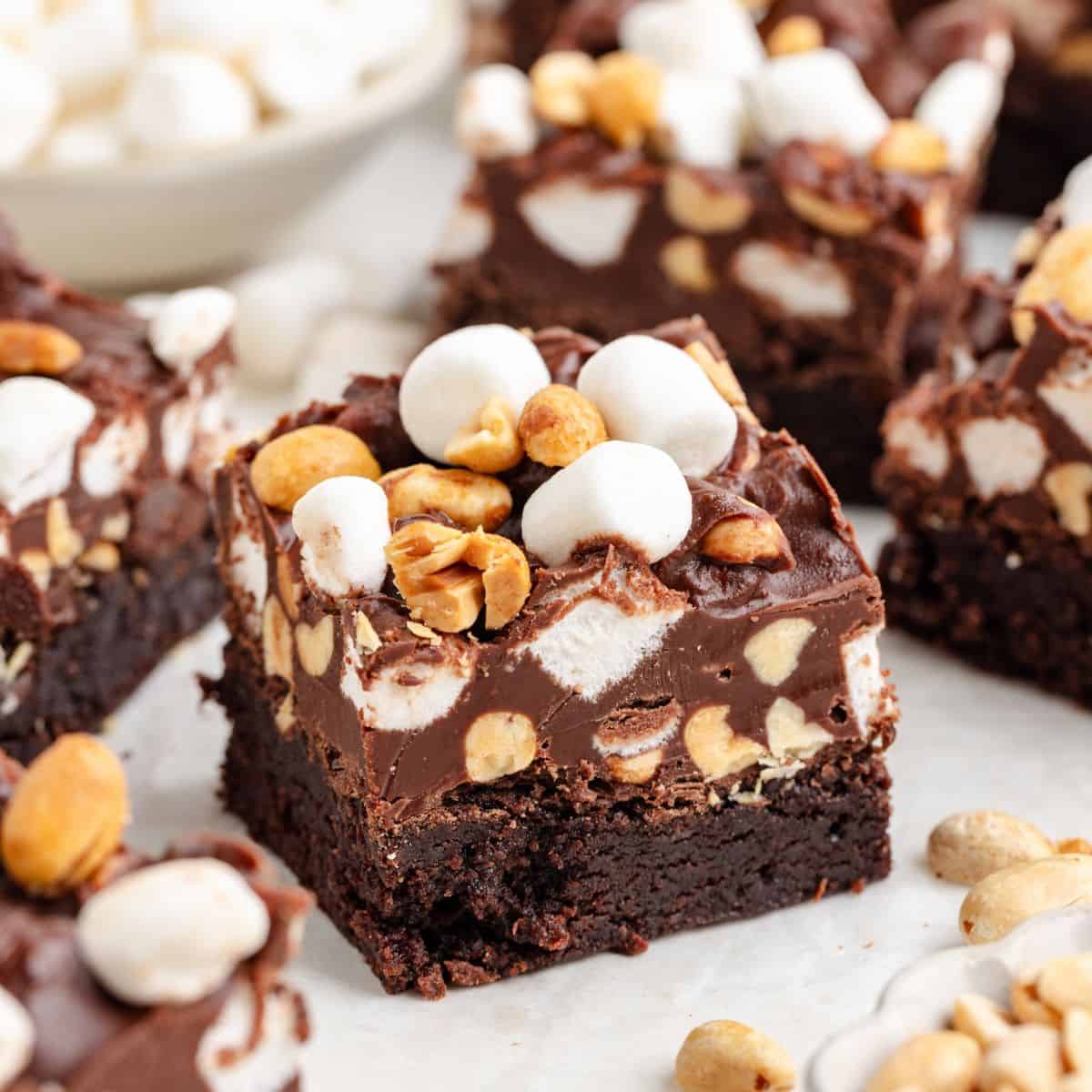 Rocky road brownies with peanuts, marshmallows, surrounded by more brownies and toppings on a white surface.