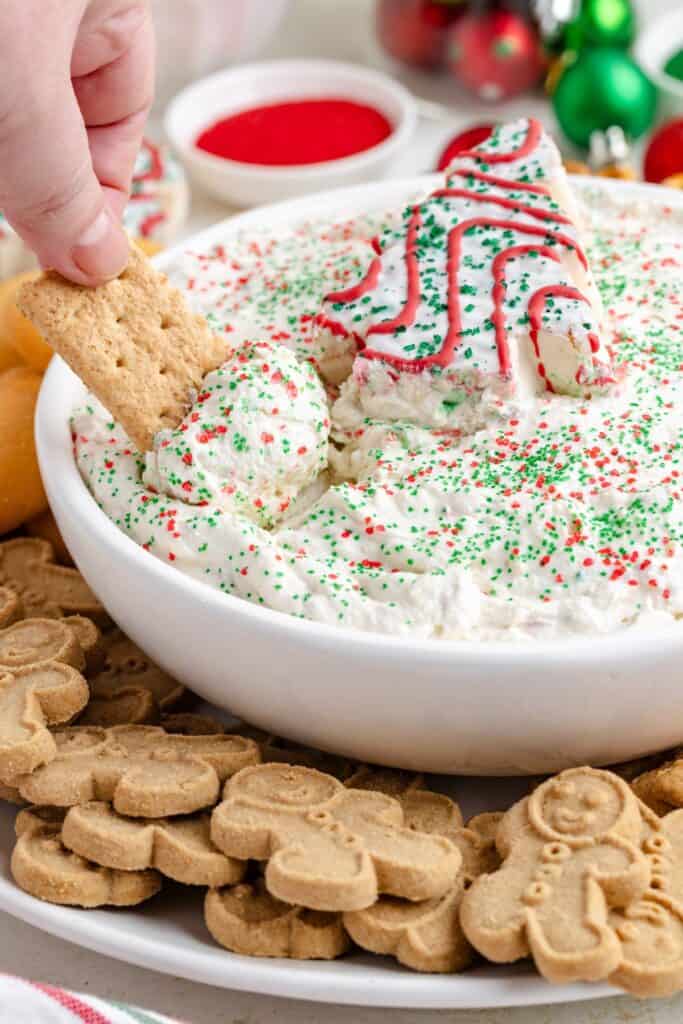 A hand dips a graham cracker into a bowl of Christmas tree cake dessert dip topped with sprinkles and a tree cookie.
