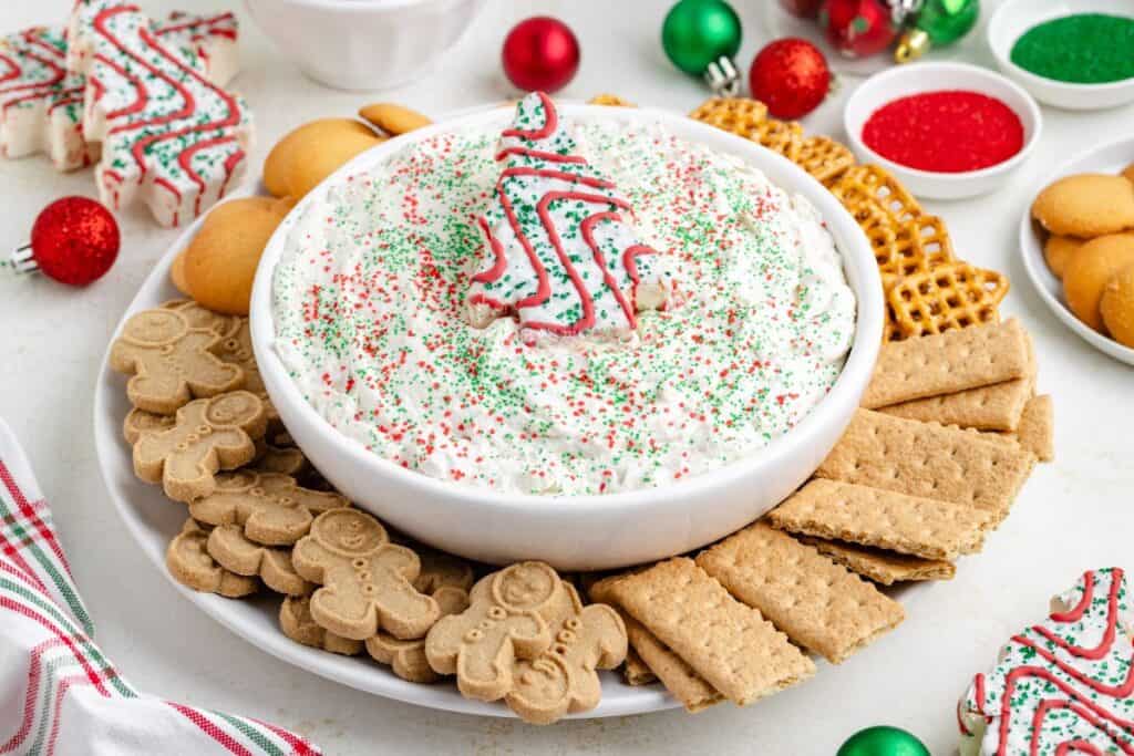 Christmas tree cake dip in a bowl with sprinkles and cookie on top, surrounded by assorted crackers and cookies.