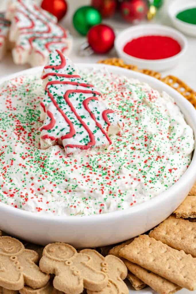 Christmas dessert dip topped with red and green sprinkles, surrounded by cookies and crackers, with a Christmas tree cookie.