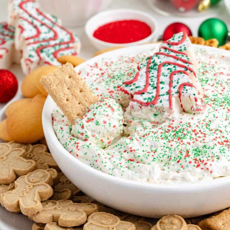 Christmas tree cake dip in a bowl topped with a Christmas tree snack cake and sprinkles, surrounded by cookies and crackers.