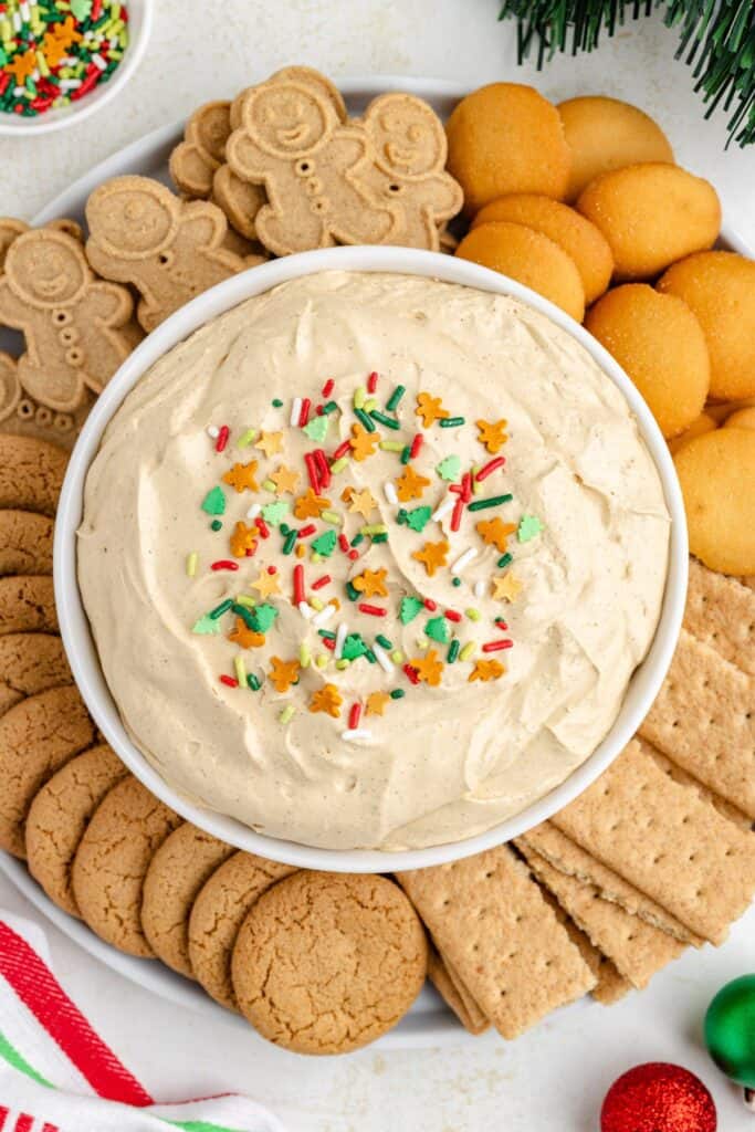 Gingerbread cream cheese dip with holiday sprinkles in a bowl, surrounded by assorted cookies and crackers on a platter.