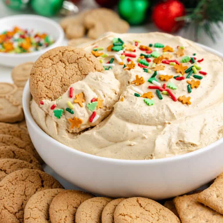 A bowl of creamy gingerbread dip with holiday sprinkles, surrounded by cookies; one cookie is dipped into the dip.