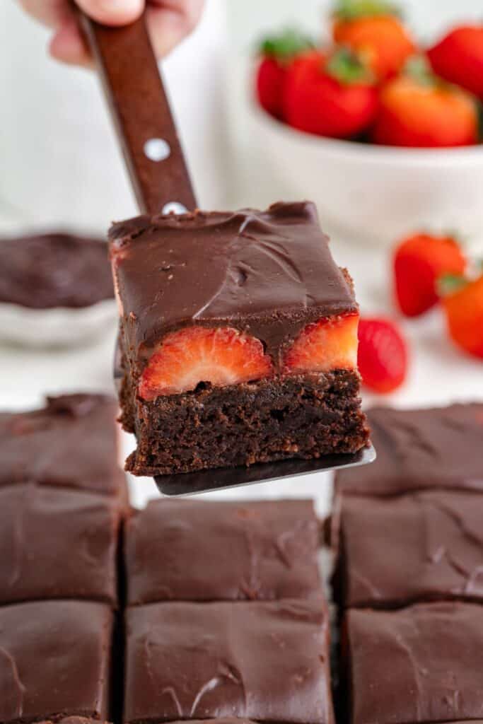 Spatula lifts a chocolate covered strawberry brownie; more brownies and strawberries behind.