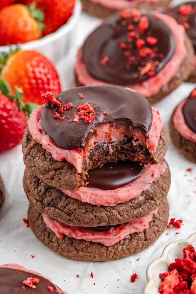 Stack of chocolate strawberry cookies with pink frosting and chocolate glaze; top cookie bitten. Fresh strawberries in background.